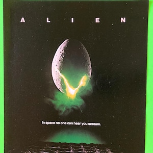 May include: A rectangular poster featuring a black background with the word ALIEN at the top. A textured, egg-shaped object is centered, emitting a bright yellow and green glow from a crack. Below, the text reads: In space no one can hear you scream.