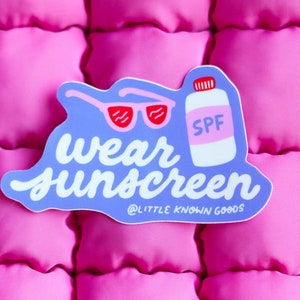 May include: A blue sticker with white text that says "wear sunscreen" and a graphic of sunglasses and a bottle of sunscreen that says "SPF". The sticker has the text "@little known goods" at the bottom.