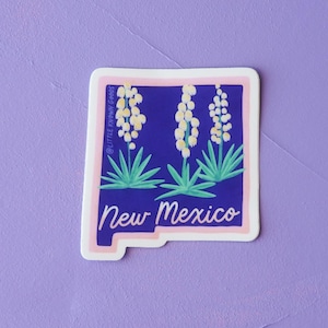 May include: A sticker featuring the outline of New Mexico in white, with a blue interior. The design includes white flowers and green leaves. The text "New Mexico" is written in cursive. The sticker has a pink border.