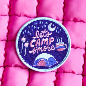 May include: A blue and pink embroidered patch with the text "Let's Camp S'more" on a dark blue background. The patch features a tent, a cup of hot chocolate, and marshmallows on sticks.