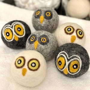 Peut inclure: Six boules de lavage en feutre en forme de hibou, en gris, blanc et noir, avec des yeux et des becs jaunes. Les boules sont sur une serviette blanche.