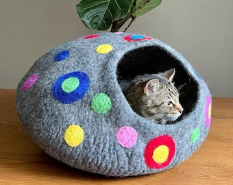 Könnte beinhalten: Ein graues Filzkatzenbett mit einem bunten Punkte-Muster. Das Bett hat eine runde Öffnung, durch die die Katze hineingehen kann. Eine getigerte Katze ruht im Bett.