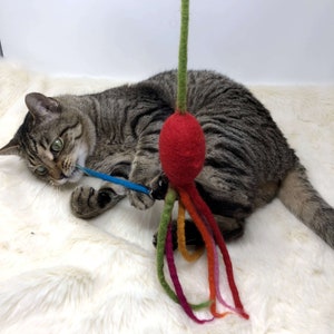 May include: A tabby cat plays with a handmade cat toy. The toy features a red felt ball with colorful felt tentacles and a green stem. The cat is lying on a white, fluffy surface, focused on the blue felt string.