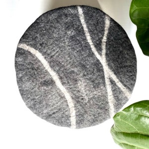 May include: A round gray felt cushion with white abstract lines. The cushion is perfect for adding a touch of modern style to any room.