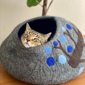 May include: A gray felt cat bed with a blue and brown tree design. A tabby cat is peeking out of the opening.
