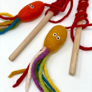 May include: Three handmade felt octopus toys. Each octopus has a colorful tail and a wooden stick handle. One octopus is orange, another is yellow, and the third is red. The tails are made of various colors, including red, blue, and green.