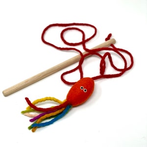 May include: A handmade cat toy featuring an orange felt octopus with rainbow tentacles and two small white eyes. The toy is attached to a wooden dowel with a red string. The toy is designed for interactive play with cats.