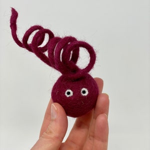 May include: A small, hand-crafted felt toy shaped like a beetroot with a curly, burgundy felt stem and two white eyes.