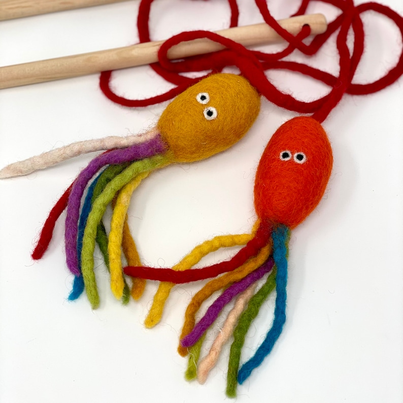 May include: Two felt octopus toys with rainbow tentacles and white eyes. One octopus is orange, the other is yellow. A red string and wooden stick are in the background. The toys are handmade.