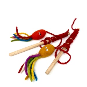 May include: Three handmade cat toys. Each toy features a felt octopus-like head in orange or yellow, with googly eyes and colorful yarn tentacles. The heads are attached to wooden dowels with red yarn.
