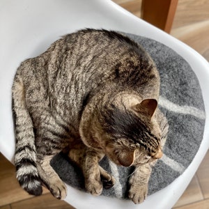 May include: A tabby cat is curled up on a gray and white patterned cat bed. The bed is on a white chair.