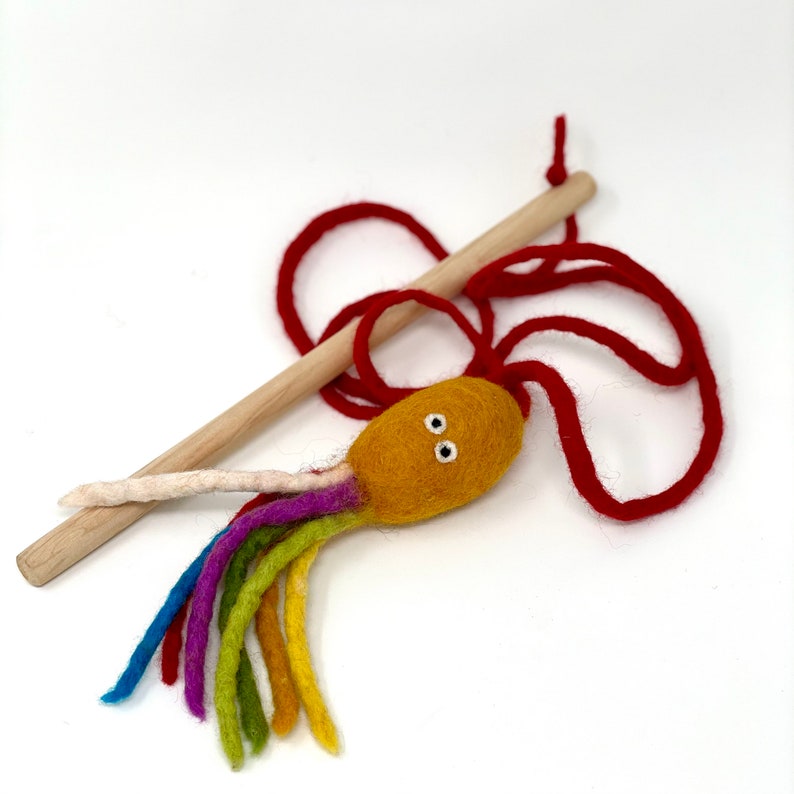 May include: A handmade cat toy featuring a yellow felt octopus with rainbow tentacles, white eyes, and a cream-colored tentacle. It has a red rope and a wooden stick. The toy is designed for interactive play.