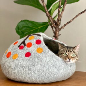 Puede incluir: Una cama para gatos de fieltro gris con una rama marrón y círculos de fieltro naranja y rojo. Un gato atigrado asoma la cabeza desde la cama.