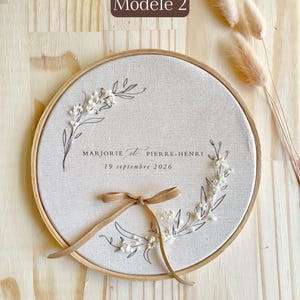 May include: A round, beige fabric embroidery hoop with a floral design and the names "MARJORIE et PIERRE-HENRI" and the date "19 septembre 2026" in black script. A tan ribbon is tied at the bottom. The text "Mod&egrave;le 2" is in a brown rectangle.