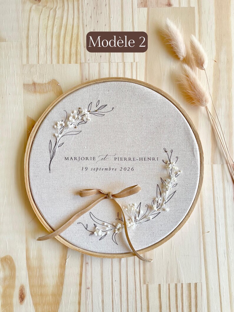 May include: A round, beige fabric embroidery hoop with a floral design and the names "MARJORIE et PIERRE-HENRI" and the date "19 septembre 2026" in black script. A tan ribbon is tied at the bottom. The text "Mod&egrave;le 2" is in a brown rectangle.