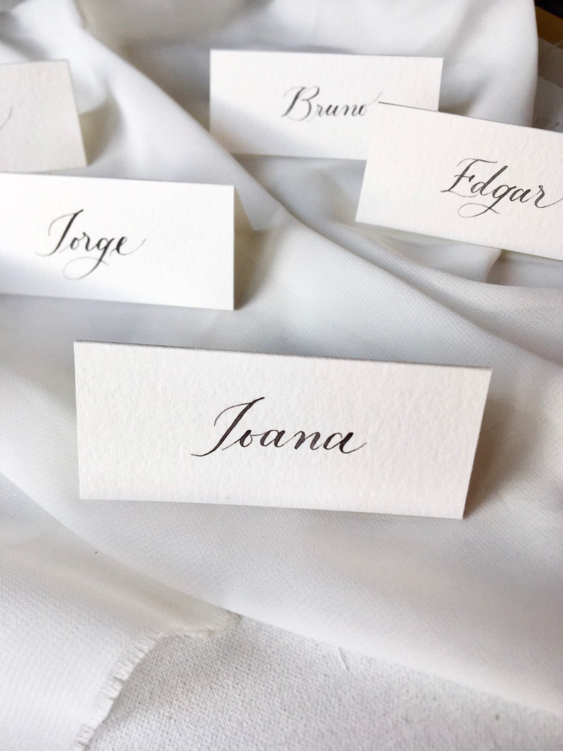 May include: Five white place cards with black calligraphy. The names on the cards are Bruno, Edgar, Jorge, Ioana, and an unreadable name.