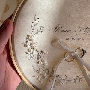 May include: A round, cream-colored fabric ring holder with a wooden hoop. The fabric is decorated with a floral design and the names "Marine & Steph" and the date "12.09.2026". Two wedding rings are attached with a cream ribbon.