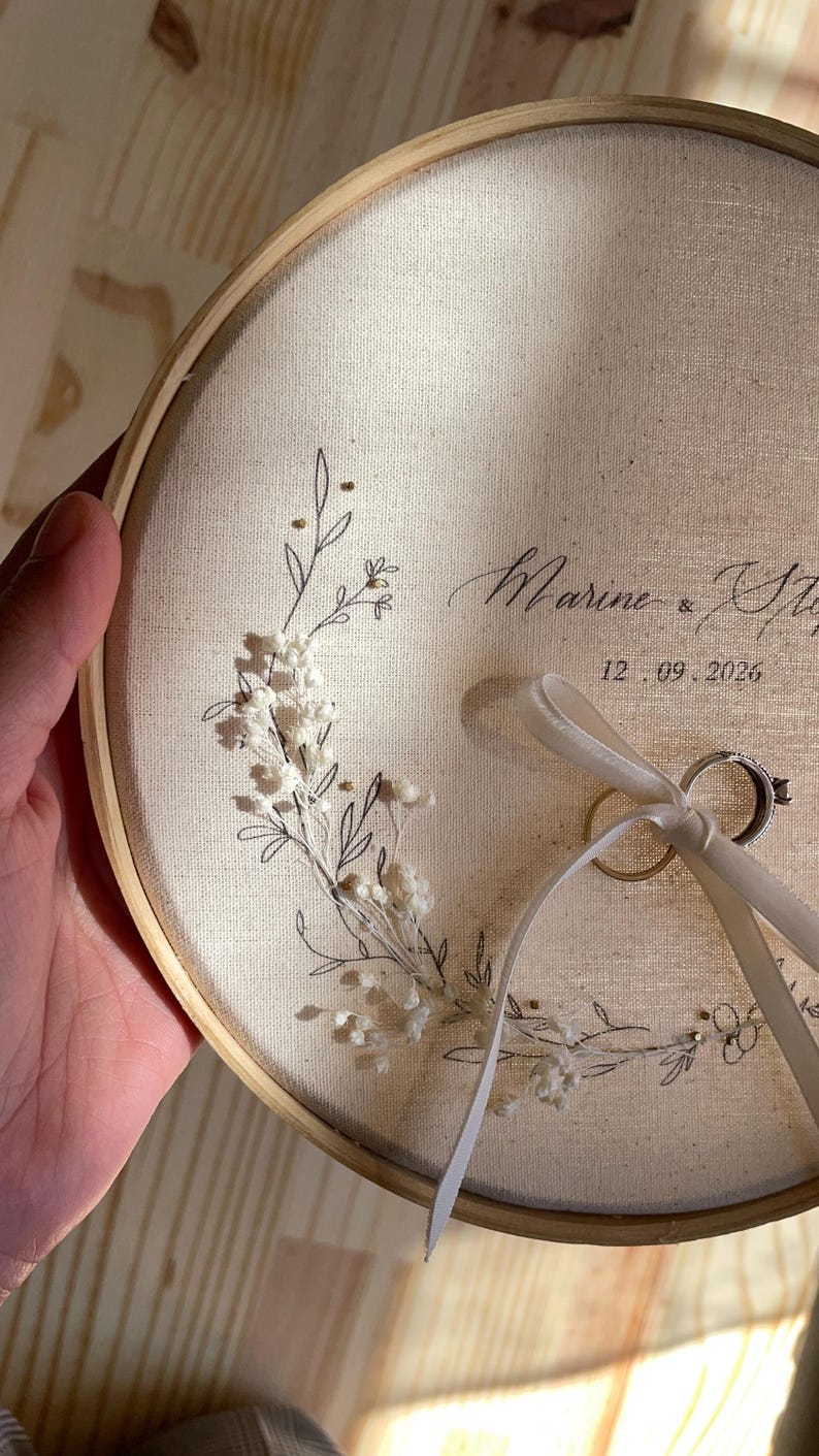 May include: A round, cream-colored fabric ring holder with a wooden hoop. The fabric is decorated with a floral design and the names "Marine & Steph" and the date "12.09.2026". Two wedding rings are attached with a cream ribbon.