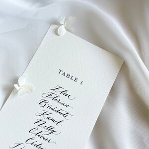 May include: A white card with black calligraphy listing the names "Elise, Florian, Benedicte, Kamel, Nelly, Olivier, Cedric" for Table 1.