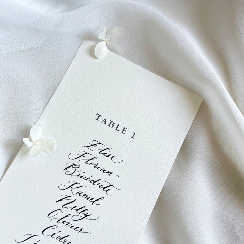 May include: A white card with black calligraphy listing the names "Elise, Florian, Benedicte, Kamel, Nelly, Olivier, Cedric" for Table 1.
