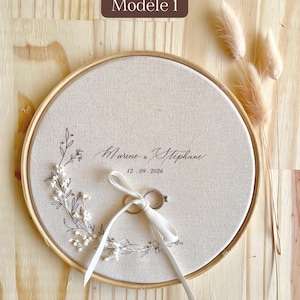 May include: A round, beige ring bearer pillow with a wooden frame. The pillow features the names "Marine & Stephane" and the date "12.09.2026" in elegant script, with a white ribbon holding two rings. Decorative floral elements and the text "Mod&egrave;le 1" are also visible.