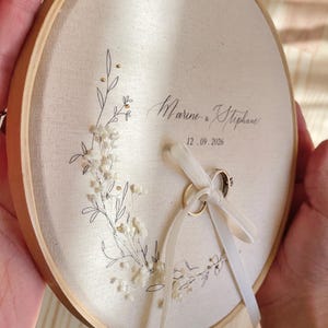 May include: A round, wooden embroidery hoop holds a cream-colored fabric with floral embroidery and the names "Marine & Stephan" and the date "12.09.2026". Two wedding rings are tied with a white ribbon. A beautiful ring bearer pillow.