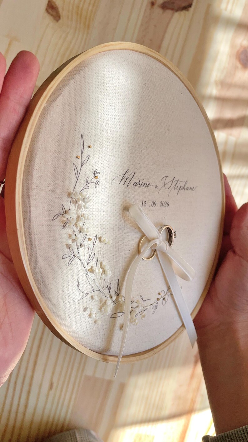May include: A round, wooden embroidery hoop holds a cream-colored fabric with floral embroidery and the names "Marine & Stephan" and the date "12.09.2026". Two wedding rings are tied with a white ribbon. A beautiful ring bearer pillow.