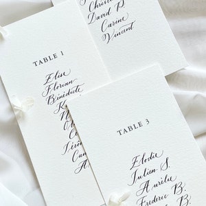 May include: Three white cards with black calligraphy listing table numbers and guest names. The cards are arranged in a staggered pattern, with the top card partially obscuring the card below it. The text on the cards reads: "TABLE 2", "TABLE 1", "TABLE 3".