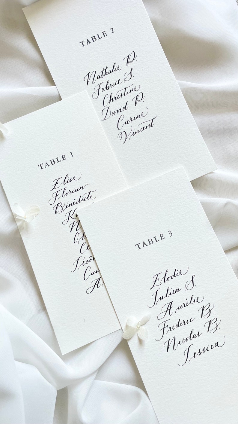 May include: Three white cards with black calligraphy listing table numbers and guest names. The cards are arranged in a staggered pattern, with the top card partially obscuring the card below it. The text on the cards reads: "TABLE 2", "TABLE 1", "TABLE 3".