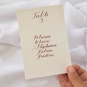 May include: A white card with the text "Table 3" written in cursive. Below the table number are the names Marin, Maia, Stephanie, Fabien, and Frederic written in cursive.