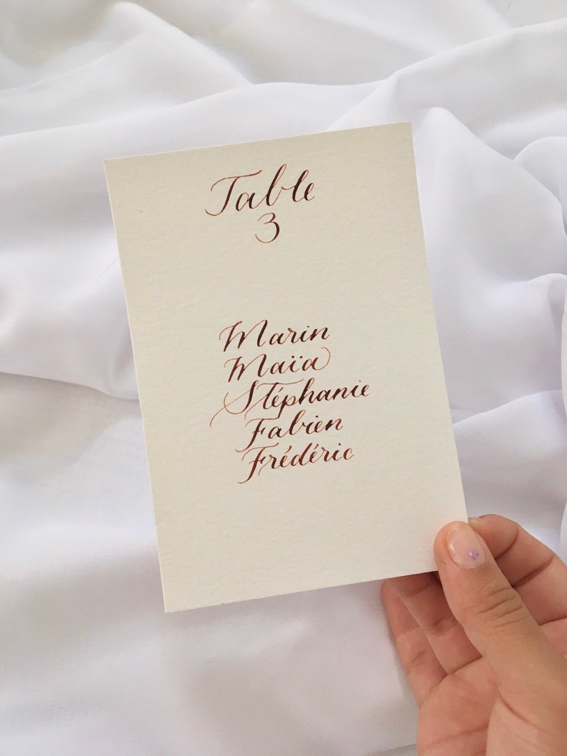 May include: A white card with the text "Table 3" written in cursive. Below the table number are the names Marin, Maia, Stephanie, Fabien, and Frederic written in cursive.