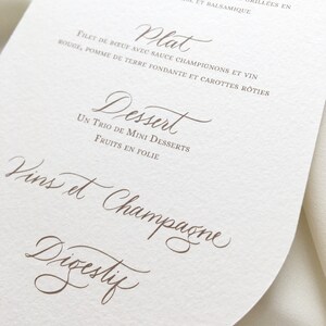 May include: A white, round menu card with a brown, handwritten menu. The menu includes the following sections: Entree, Plat, Dessert, Vins et Champagne, and Digestif.