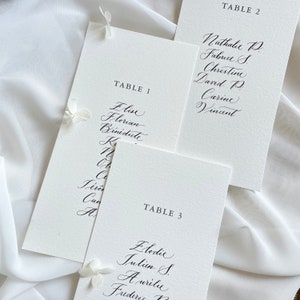 May include: Three white cards with black calligraphy listing table numbers and guest names. The cards are labelled "Table 1", "Table 2", and "Table 3".