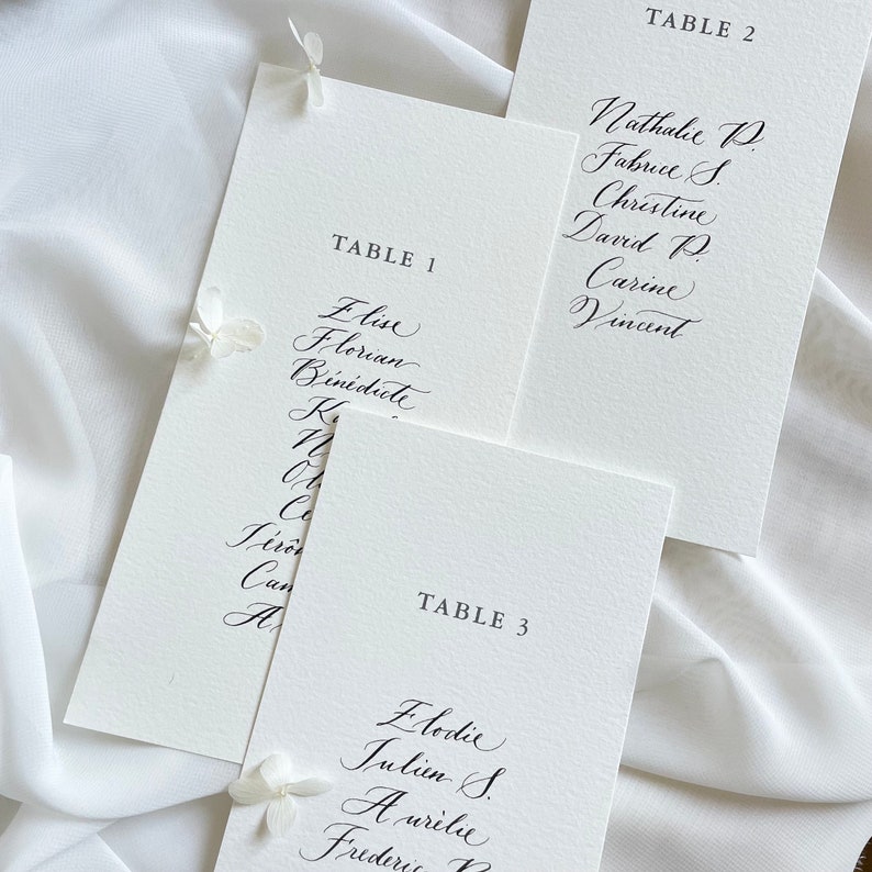 May include: Three white cards with black calligraphy listing table numbers and guest names. The cards are labelled "Table 1", "Table 2", and "Table 3".
