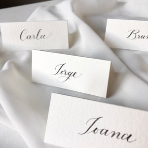 May include: Four white place cards with black calligraphy. The names on the cards are Carla, Jorge, Bruna, and Joana.