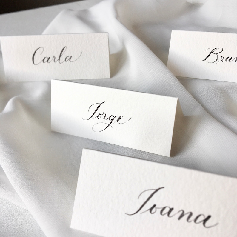 May include: Four white place cards with black calligraphy. The names on the cards are Carla, Jorge, Bruna, and Joana.