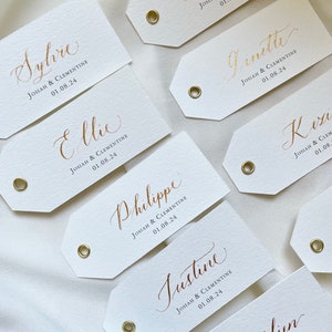 K&ouml;nnte beinhalten: Wei&szlig;e rechteckige Etiketten mit goldenen Kalligraphie-Namen und dem Text "Josiah & Clementine 01.08.24" auf jedem Etikett.