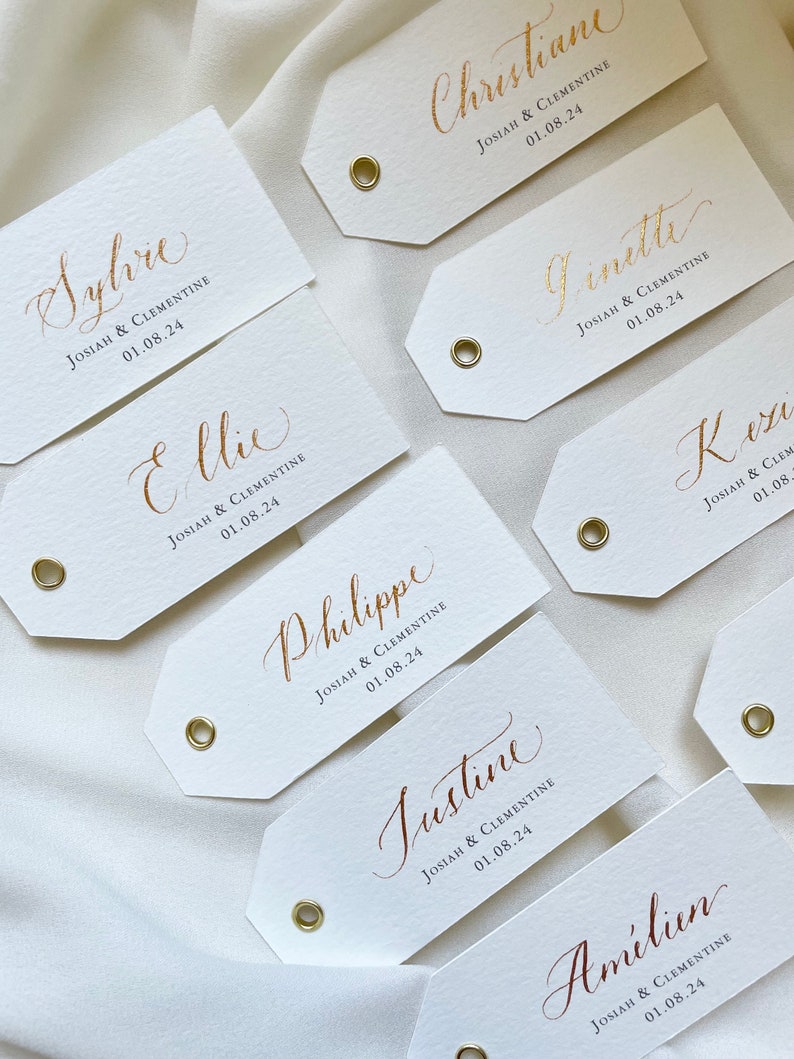 K&ouml;nnte beinhalten: Wei&szlig;e rechteckige Etiketten mit goldenen Kalligraphie-Namen und dem Text "Josiah & Clementine 01.08.24" auf jedem Etikett.