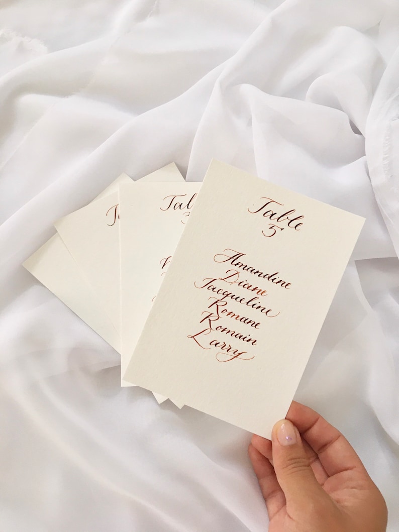 May include: A white card with the text "Table 5" written in cursive script. Below the table number are the names Alexandrine, Diane, Jacqueline, Romain, Romain, and Larry, also written in cursive script.