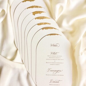 May include: A stack of oval-shaped white cards with gold accents. The cards have a menu printed on them in French. The menu includes the following items: Plat, Fromages, Dessert.