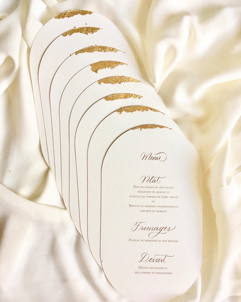 May include: A stack of oval-shaped white cards with gold accents. The cards have a menu printed on them in French. The menu includes the following items: Plat, Fromages, Dessert.