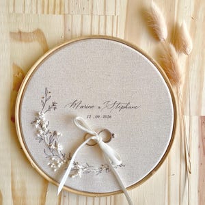 May include: A round, beige fabric ring holder with a wooden frame. The fabric is embroidered with floral designs and the names "Marine & Stephane" and the date "12.09.2026". Two rings are tied with a white ribbon. Dried flowers are in the background.