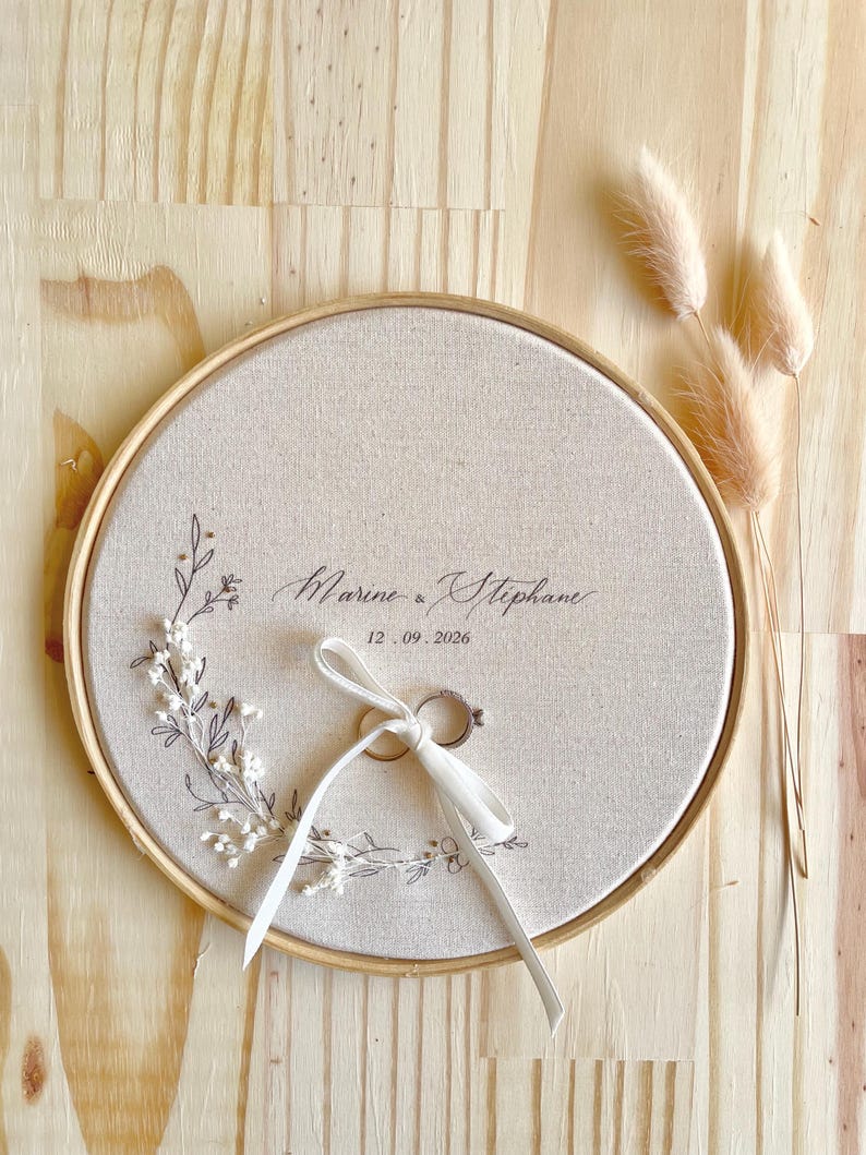 May include: A round, beige fabric ring holder with a wooden frame. The fabric is embroidered with floral designs and the names "Marine & Stephane" and the date "12.09.2026". Two rings are tied with a white ribbon. Dried flowers are in the background.