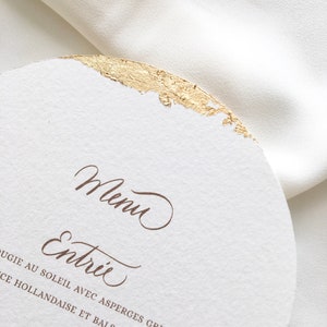 May include: A white card with gold foil accents and brown text. The text reads "Menu" and "Entree" followed by "Asparagus grilled in butter with hollandaise and balsamic".