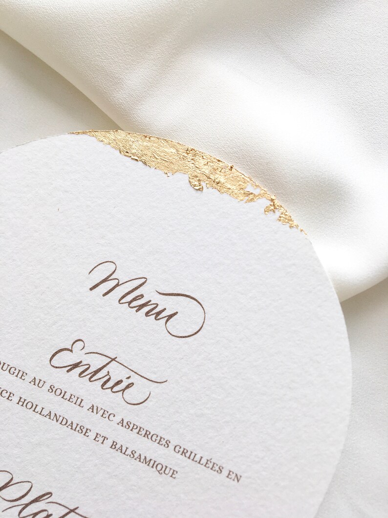 May include: A white card with gold foil accents and brown text. The text reads "Menu" and "Entree" followed by "Asparagus grilled in butter with hollandaise and balsamic".