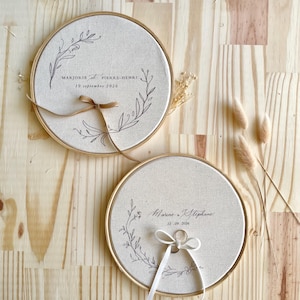 May include: Two round, beige wedding ring holders with gold-colored frames. Each holder has a floral design and text. One has a brown ribbon, the other a white ribbon with a ring. The background is a light wood surface.