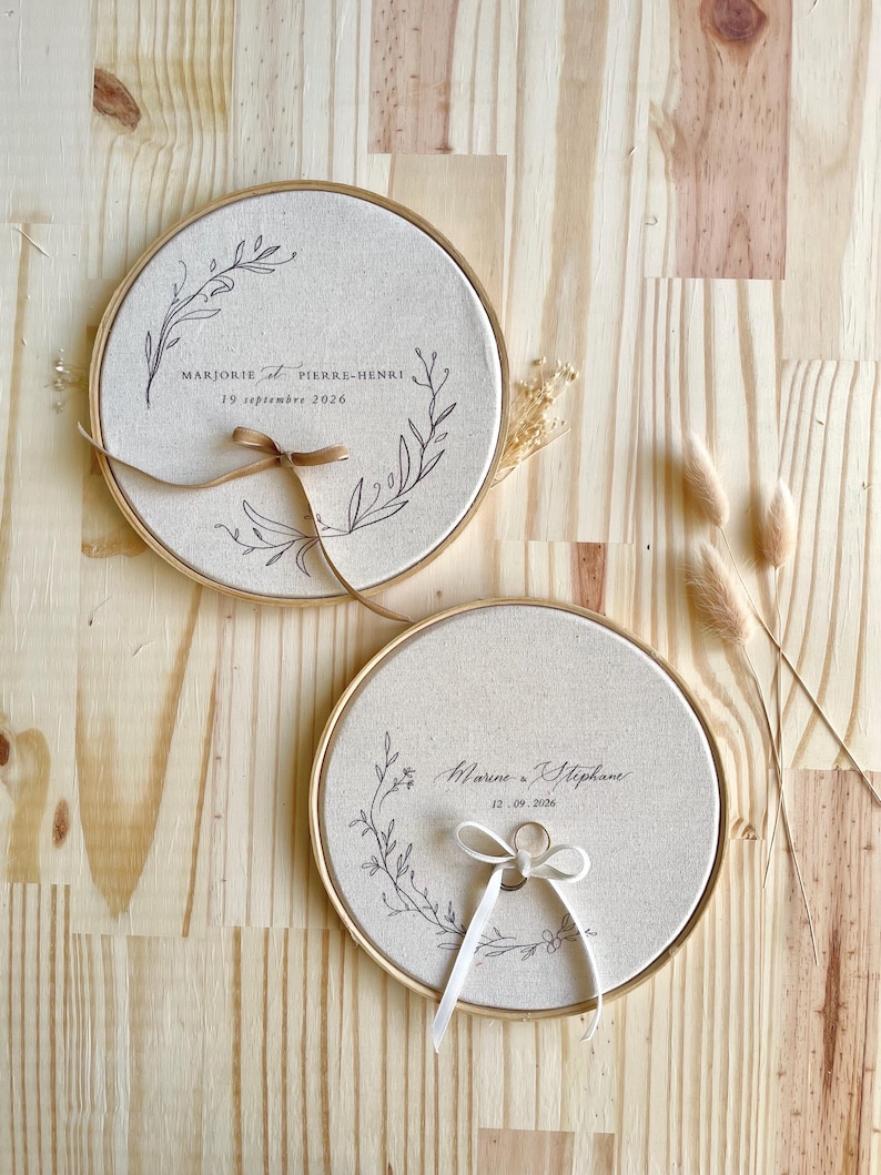 May include: Two round, beige wedding ring holders with gold-colored frames. Each holder has a floral design and text. One has a brown ribbon, the other a white ribbon with a ring. The background is a light wood surface.