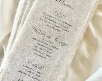 Può includere: Un menu in lino rustico con un menu scritto a mano in inchiostro nero. Il menu include una selezione di piatti, tra cui un Cote de Boeuf, un piatto di formaggi, dessert e bevande. Il menu è scritto in francese.