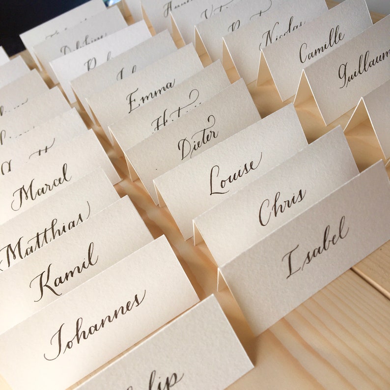 May include: A set of place cards with names written in calligraphy. The cards are folded in half and arranged in rows on a wooden surface.