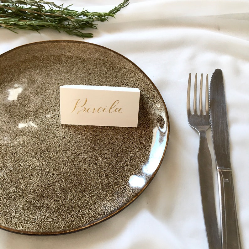 May include: A white place card with the name "Priscilla" written in gold calligraphy on a brown speckled plate with a silver fork and knife on a white tablecloth.